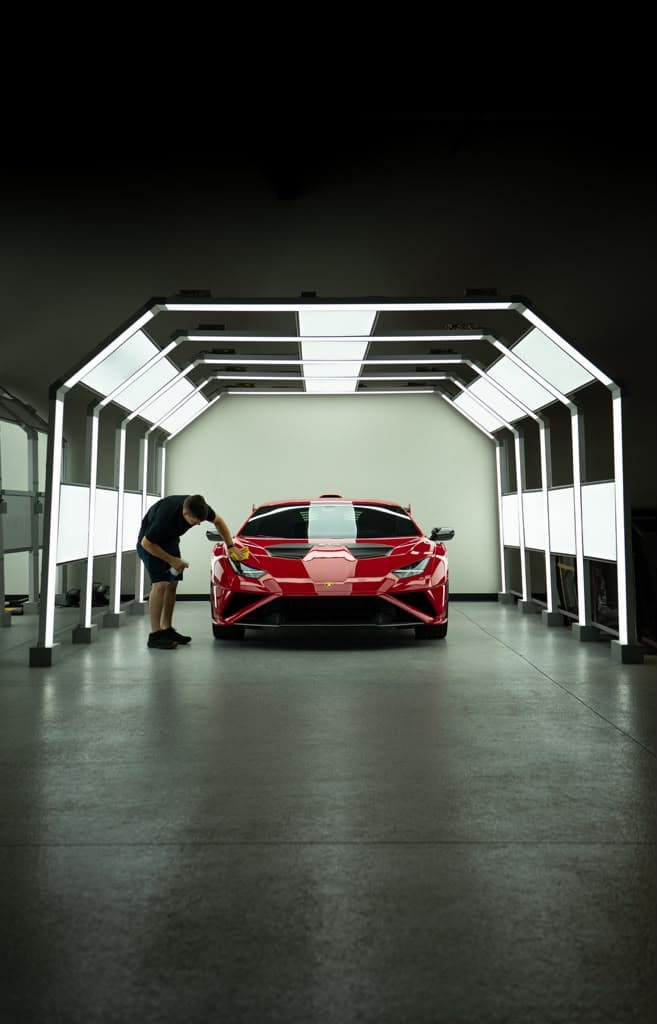 A detailer leaning over the hood of a red supercar under the light bars of the detail bay