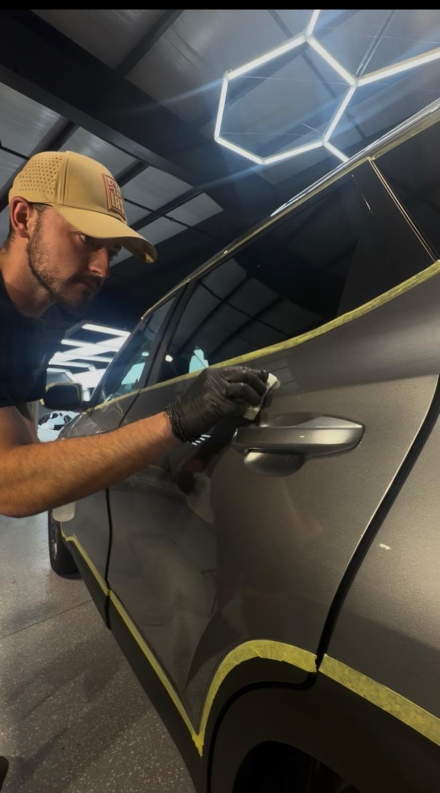 A detailer in a shop cap wiping Formula 1051 onto a car door panel by hand under the bay's hex light